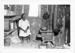 Myrtle Lawrence's neighbor.  Young woman sits near the wood stove and cooking pots in substandard living conditions.