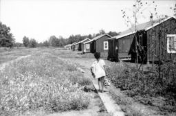 Young Black girl walks down a dirt path carrying a pail toward new houses