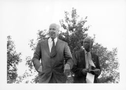 Norman Thomas speaking at an outdoor STFU meeting. An unidentified Black man stands with him on stage