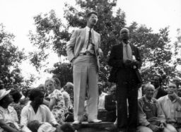 Two speakers standing on a platform at an outdoor STFU meeting, Sylvia Lawrence seated amongst the listeners