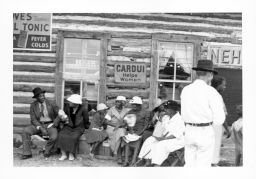 People dressed in best clothes for a STFU meeting sit outside a store. Behind them, a sign advertises Cardui tonic for women and other remedies for sickness