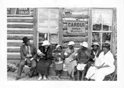 A group of women with one man and one child, all dressed in best clothes for a STFU meeting sit outside a store.  Behind them, a sign advertises Cardui tonic for women