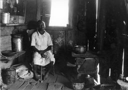 Young woman sits near the wood stove and  cooking pots in substandard living conditions. From the image: "Neighbor of the Lawrences," "Daylight"