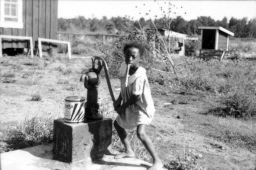 A young Black girl operates a water pump at Delta Cooperative with balloon in her mouth