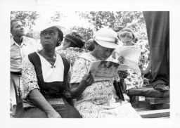 Photo verso reads "Union meeting - reading is not easy."  An unidentified woman and Sylvia Lawrence read the "Sharecroppers' Voice" during an outdoor STFU meeting