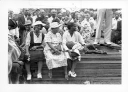 Group of Blacks and Whites standing around and sitting on the platform during an outdoor STFU meeting