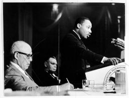 Martin Luther King, Jr., giving a speech, while George Meany, also at the speakers' table, listens