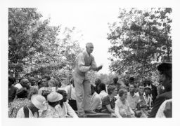 Christian minister speaking at an outdoor STFU meeting with listeners seated around the base of the platform