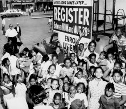A large group of African American children gather around a sign encouraging people to register to vote.