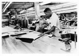 An African American man cuts fabric in a large garment shop with a power cutter.