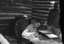 Man seated at a table writes in book while another man waits