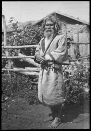 An elderly Ainu man, in front of thatched houses