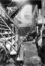 "13. 37 Ridge St. Yard with closets." Alley with doorways, barrel, piles of cloth scraps and stairs leading to landings. Next yard has laundry lines hanging over the alley.