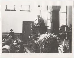 Wideshot of Paul Robeson speaking at wooden podium at Lorraine Hansberry's funeral