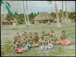 View of several young boys, seated, in front of thatched buildings, surrounded by palm trees, probably in Samoa
