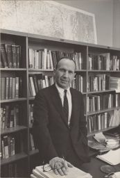 Portrait photograph of Clinton Rossiter, Professor of Government, standing in his office with his hand on a stack of texts.