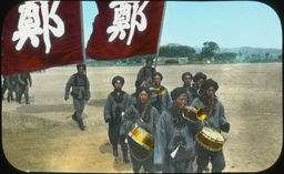 A musical band of Japanese troops, some carrying flags, process across a field