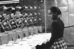 A young African American woman watches at least seven sewing machines working simultaneously.