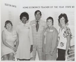 Four women and one man in the middle in front of Home Economics Teacher of the Year sign