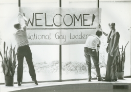 Three men hanging up a Welcome National Gay Leaders banner