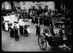 Women in horse-drawn carriage and on foot march in street for voting rights carrying banners "Mothers Prepare the Children for the World..." "Women Need Votes..." and "Suffrage Pioneers..."