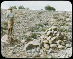 Young man in breeches, standing near a rock pile (potential burial)