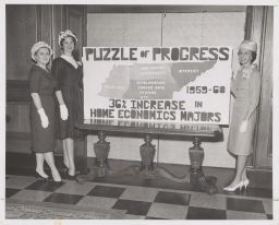 Three women with Puzzle of Progress poster