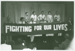 Richard Berkowitz and ten other men reading the Denver Principles behind a "Fighting For Our Lives" banner at the National Lesbian and Gay Health conference
