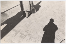 Self-portrait for photo class assignment, 1974. Her shadow following a man with a wrinkled coat and a cardboard box on Market St., San Francisco, near her home. "The transients on Market Street were my friends. The area was my home. I took my pictures there."