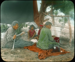 Two native women weave under a tree, while an infant looks on
