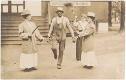 Three women holding sticks and brooms surrounding an African-American man